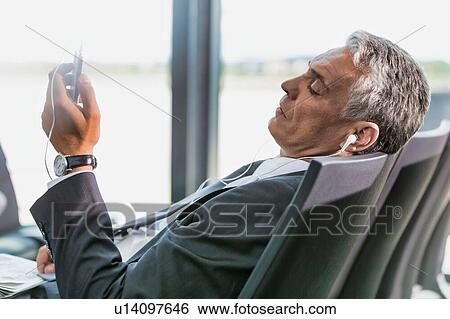 Portrait of mature businessman listening to music on his smartphone while waiting for boarding in airport View Large Photo Image Stock Photograph - Portrait of mature businessman listening to music on his smartphone while waiting for boarding in airport. Fotosearch