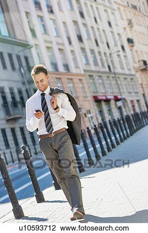 Stock Image - Portrait of mature businessman using smartphone while walking on pavement after work. Fotosearch
