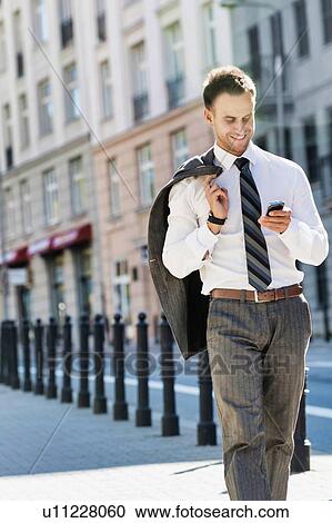 Stock Image - Portrait of mature businessman using smartphone while walking on pavement after work. Fotosearch
