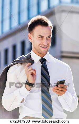 Stock Image - Portrait of mature businessman using smartphone while walking on pavement after work. Fotosearch