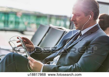 Stock Photo - Portrait of mature businessman using digital tablet with earphones on while waiting for boarding in airport. Fotosearch