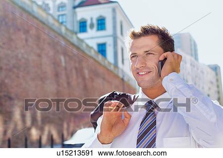 Stock Image - Portrait of mature confident businessman smiling and talking on smartphone while walking on pavement. Fotosearch