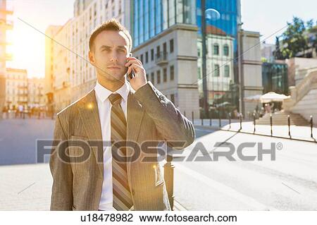 Stock Image - Portrait of mature confident businessman smiling and talking on smartphone while walking on pavement. Fotosearch