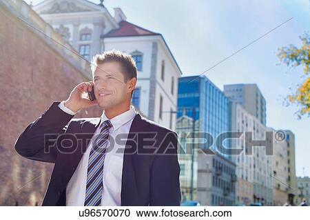 Stock Image - Portrait of mature confident businessman smiling and talking on smartphone while walking on pavement. Fotosearch