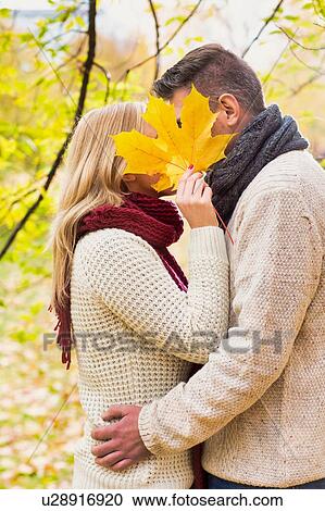 Stock Image - Portrait of Mature couple kissing in park while the woman is covering their face with maple leaf. Fotosearch