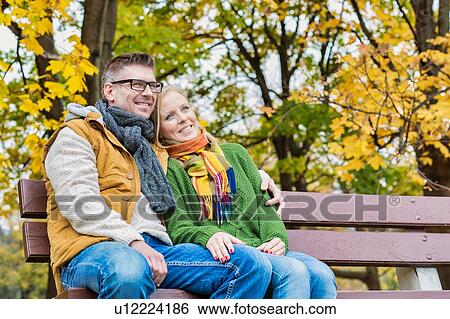 Stock Photograph - Portrait of mature couple sitting on bench while enjoying autumn in park. Fotosearch