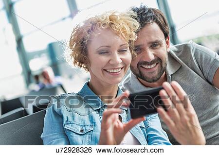 Stock Photograph - Portrait of mature couple taking and looking at their selfie while waiting for boarding in airport. Fotosearch
