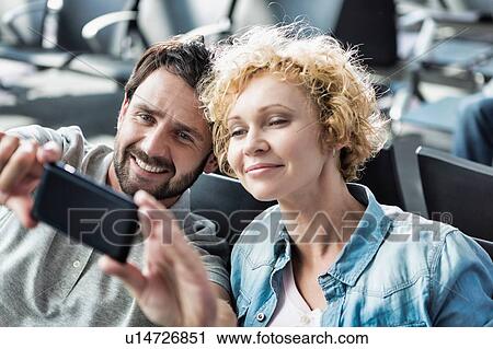 Stock Image - Portrait of mature couple taking selfie while waiting for boarding in airport. Fotosearch