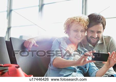Stock Photograph - Portrait of mature couple taking selfie while waiting for boarding in airport. Fotosearch