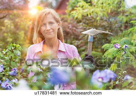 Stock Image - Portrait of mature female gardener holding fork rake. Fotosearch