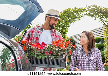 Picture - Portrait of mature gardener putting flowers on crate in car trunk while talking to woman in garden shop. Fotosearch