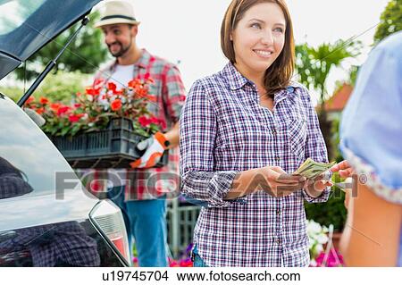 Portrait of mature gardener putting flowers on crate in car trunk while woman buyer giving cash payment View Large Photo Image Picture - Portrait of mature gardener putting flowers on crate in car trunk while woman buyer giving cash payment. Fotosearch