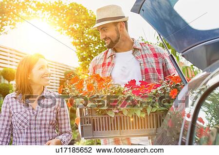 Stock Image - Portrait of mature gardener putting flowers on car trunk while talking to woman in garden shop. Fotosearch
