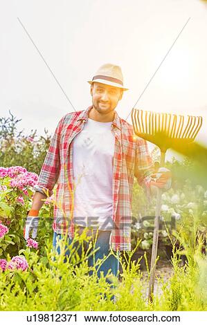 Stock Image - Portrait of mature gardener standing while holding fork rake . Fotosearch