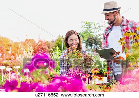 Stock Image - Portrait of mature gardener writing report on clipboard over flowers in shop with woman buyer. Fotosearch