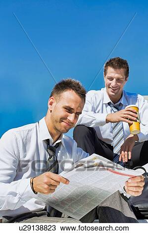 Stock Image - Portrait of mature handsome businessman reading newspaper against collegue during break. Fotosearch