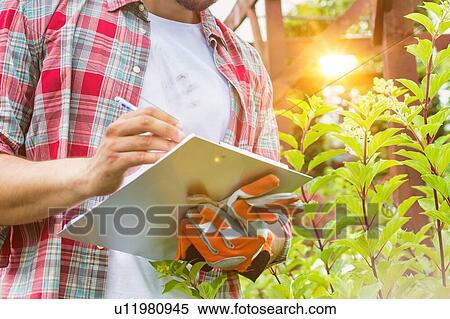 Stock Photography - Portrait of mature male gardener writing report on clipboard over flowers in shop. Fotosearch