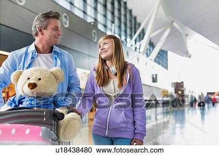 Stock Image - Portrait of mature man pushing baggage cart while walking with her daughter in airport with lens flare. Fotosearch