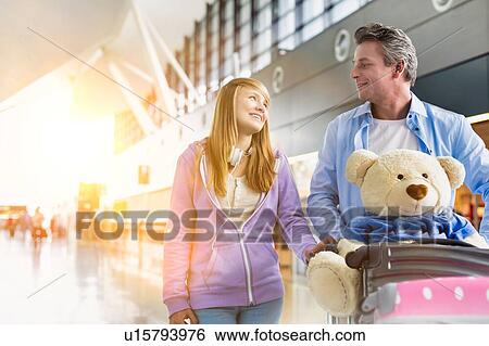 Stock Photograph - Portrait of mature man pushing baggage cart while walking with her daughter in airport with lens flare. Fotosearch