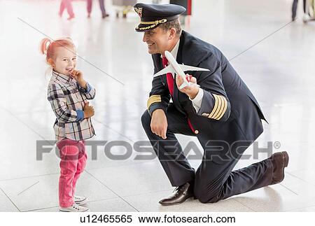 Stock Photography - Portrait of mature pilot holding airplane toy while playing with cute little girl in airport. Fotosearch