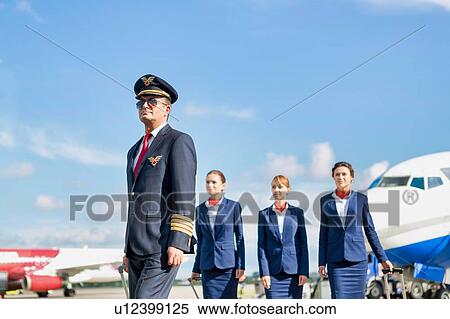 Stock Photography - Portrait of mature pilot walking with three young beautiful flight attendants against airplane in airport. Fotosearch