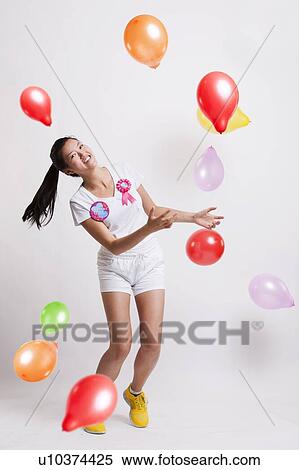 Stock Photography - Portrait of playful young woman with multicolored balloons celebrating her birthday against white background. Fotosearch