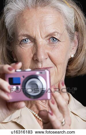 Stock Image - Portrait of senior woman holding camera against black background. Fotosearch