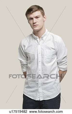 Stock Image - Portrait of serious young man with hands behind back against gray background. Fotosearch