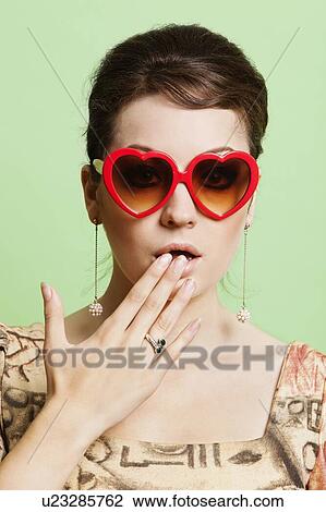 Portrait of shocked young woman wearing heart shaped sunglasses against green background View Large Photo Image Stock Image - Portrait of shocked young woman wearing heart shaped sunglasses against green background. Fotosearch