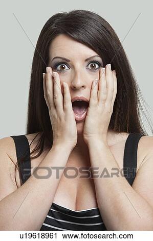Stock Image - Portrait of shocked young woman with hands on face against gray background. Fotosearch