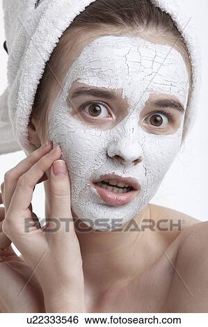 Stock Photograph - Portrait of shocked young woman with face mask peeling off against white background. Fotosearch