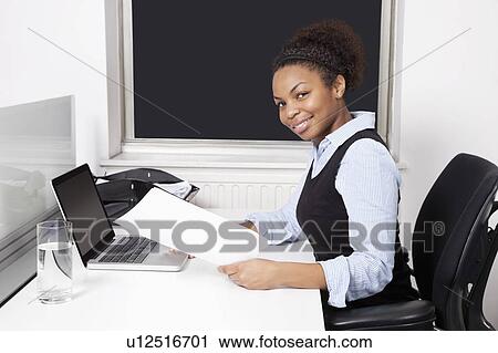 Stock Image - Portrait of smiling businesswoman with document using laptop at desk in office. Fotosearch