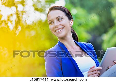 Stock Image - Portrait of Smiling young attractive businesswoman holding digital tablet while standing in park. Fotosearch