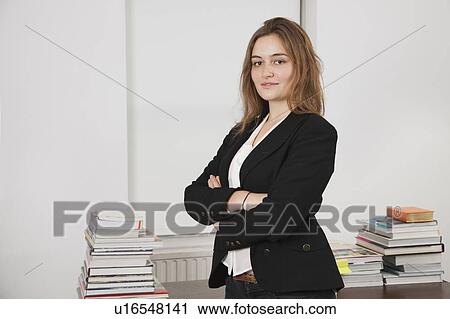 Stock Image - Portrait of smiling young businesswoman with stack of books on desk in office. Fotosearch