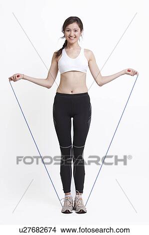 Picture - Portrait of smiling young woman exercising with skipping rope against white background. Fotosearch
