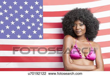 Stock Image - Portrait of stylish young woman in bikini standing against American flag. Fotosearch