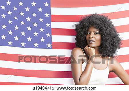 Stock Photography - Portrait of stylish young woman with fizzy hairstyle standing against American flag. Fotosearch