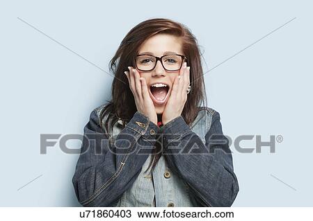 Stock Image - Portrait of surprised young woman with hands on cheeks against light blue background. Fotosearch