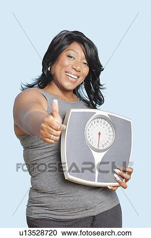 Stock Image - Portrait of woman giving thumbs up while holding scales over blue background. Fotosearch