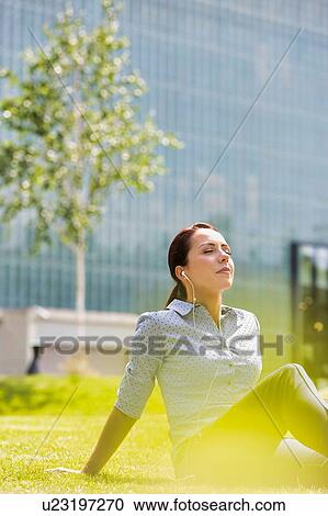 Stock Image - Portrait of young arrractive businesswoman sitting on the grass while listening on music with earphones on . Fotosearch