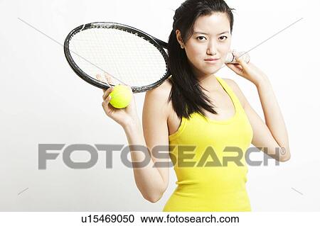 Stock Image - Portrait of young Asian female tennis player holding racket and ball against white background. Fotosearch