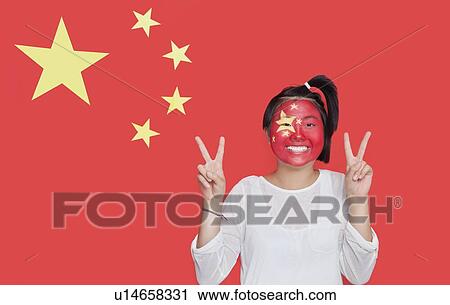 Stock Image - Portrait of young Asian woman with painted face gesturing peace sign against Chinese flag. Fotosearch
