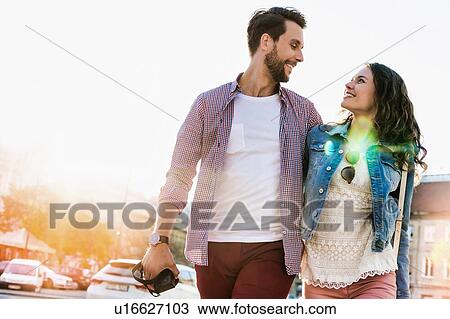 Stock Image - Portrait of young attractive couple walking in the middle of the city with lens flare in background. Fotosearch