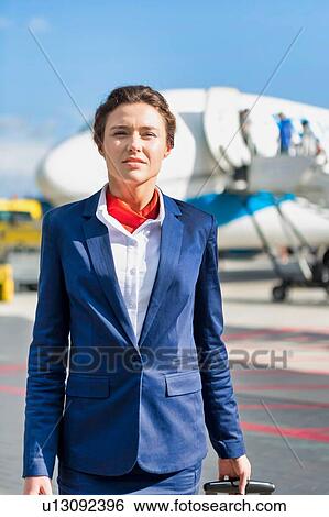 Stock Photograph - Portrait of young attractive flight attendant standing against airplane in airport. Fotosearch