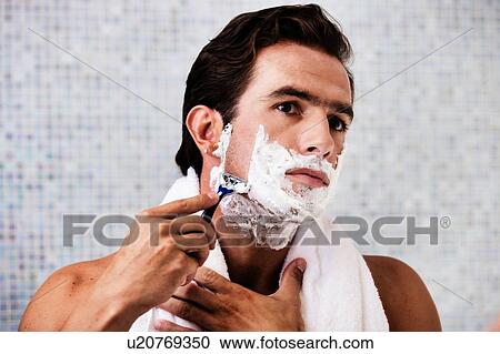 Stock Image - Portrait of young attractive man applying beard foam and shaving his beard while looking in the mirror. Fotosearch