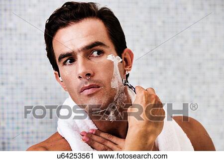 Stock Image - Portrait of young attractive man applying beard foam and shaving his beard while looking in the mirror. Fotosearch