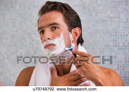 Stock Image - Portrait of young attractive man shaving his beard while looking in the mirror. Fotosearch