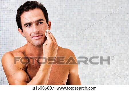 Stock Photo - Portrait of young attractive man cleansing his face with cotton and toner in the bathroom. Fotosearch