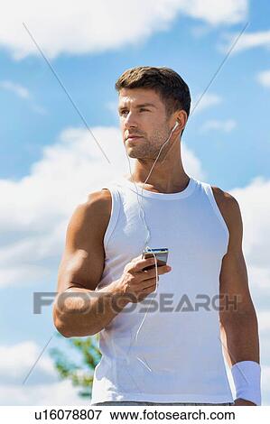 Stock Photo - Portrait of young attractive man standing while using and listening to music on his smartphone in park. Fotosearch