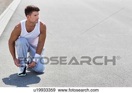 Stock Photo - Portrait of young attractive man wearing sports wear tying his rubber shoes in park. Fotosearch